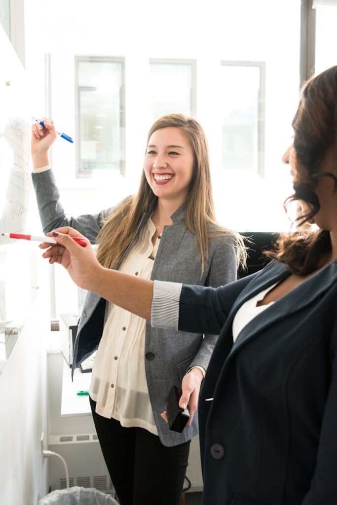Two professional women discussing ideas on a whiteboard in a modern office setting.